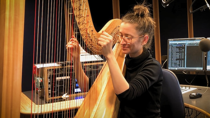 A woman plays the harp while sitting at the screens and dials of a radio console.
