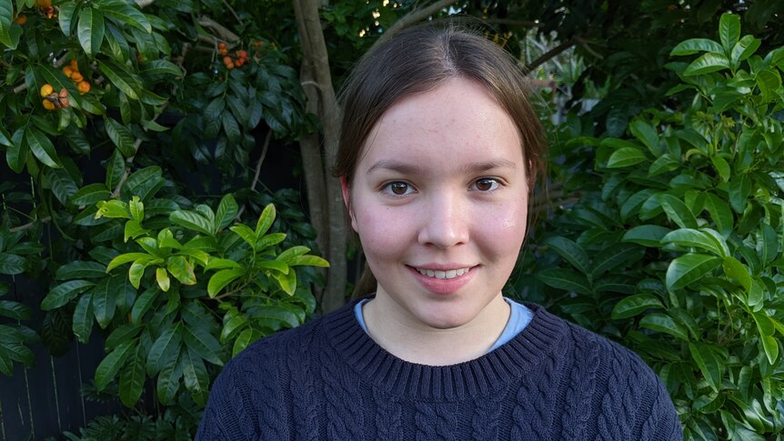 A profile image of Matilda Hockey smiling, wearing a blue sweater, brown hair tied back, in front of a green leafy bushes,