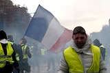 A demonstrator wearing a yellow vest grimaces through tear gas walking on wide street with a French flag in the background
