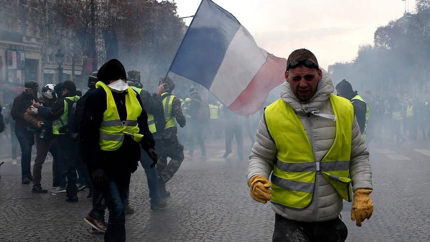 A demonstrator wearing a yellow vest grimaces through tear gas walking on wide street with a French flag in the background