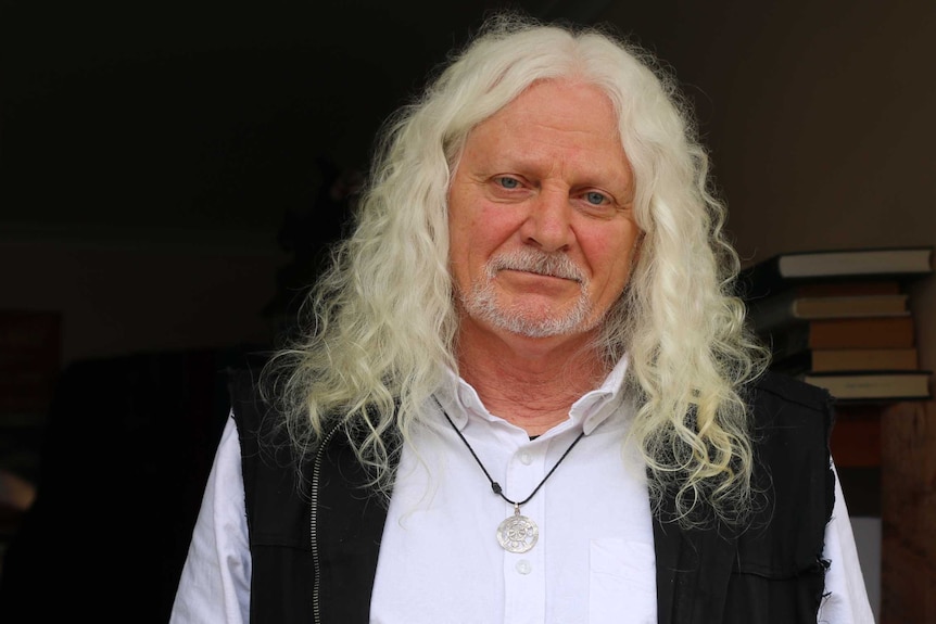 Midshot portrait of of a man with long white curly hair standing at a door entrance.