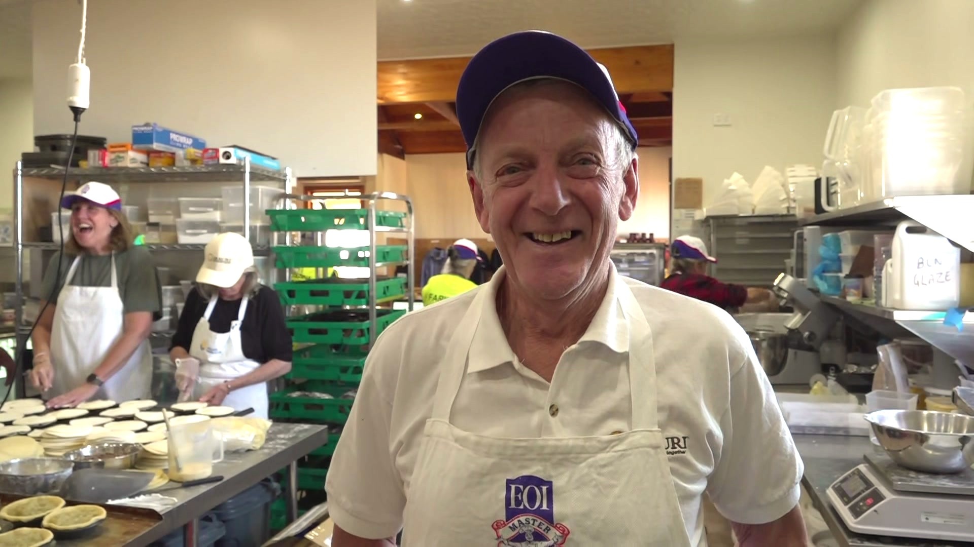 A man in a cap laughing in a kitchen with women laughing behind him.