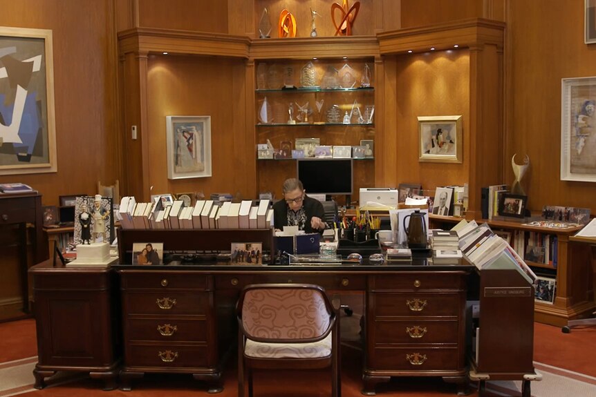 Colour still from 2018 documentary RBG of Justice Ruth Bader Ginsburg working in an office with filled with books and awards.
