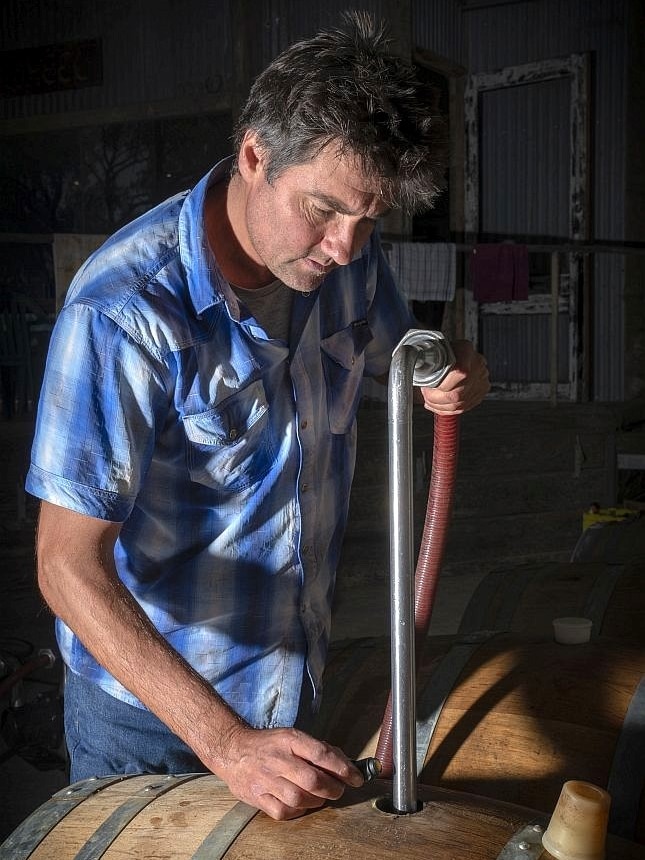 A man in a blue check shirt holds a pipe and hose placed in a hole in a wine barrel.