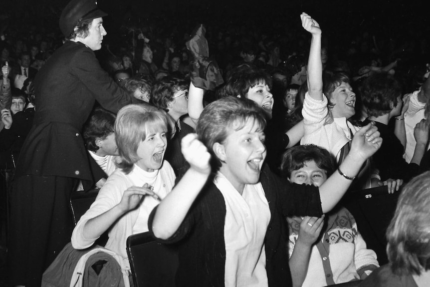 A black and white image of young women in 1960s dress cheering and screaming.