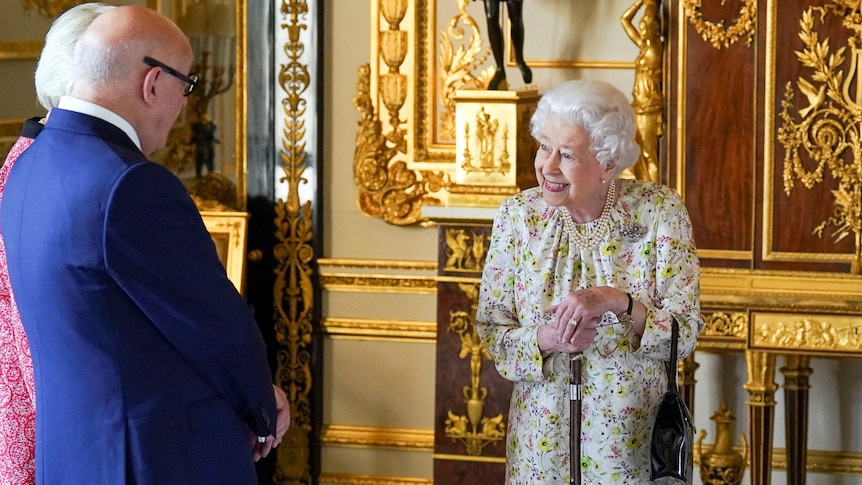 Queen Elizabeth II smiles in a floral dress as she leans on a walking cane.