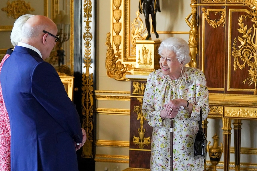 Queen Elizabeth II smiles in a floral dress as she leans on a walking cane.