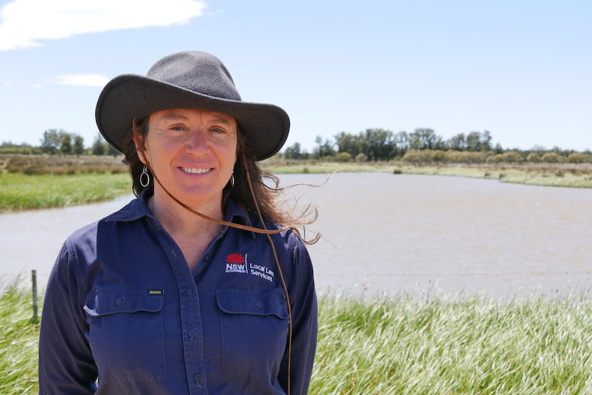 A smiling woman in a hat and a dark shirt stands in front of a wetland.