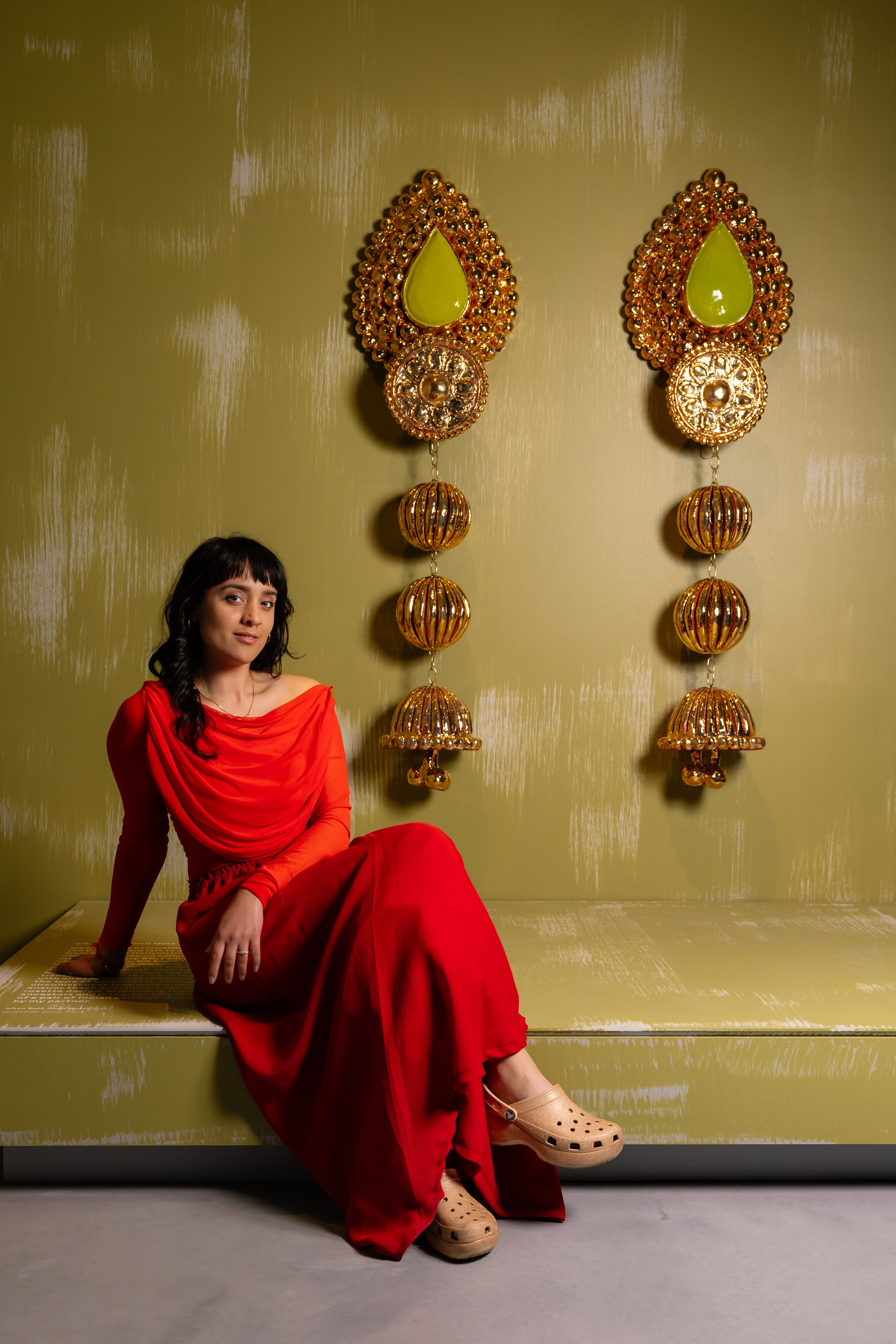 Monica Rani Rudhar wears a striking red dress and poses next to a pair of her large gold earrings in a gallery