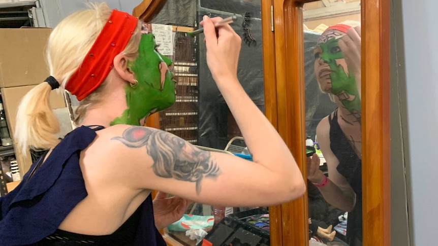 A blonde woman paints her face green in front of a mirror