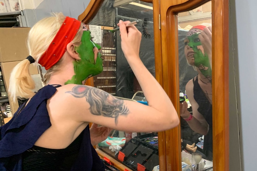 A blonde woman paints her face green in front of a mirror