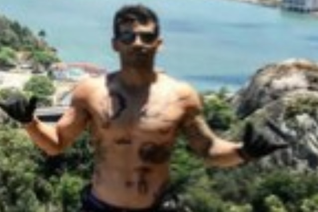 A young man, shirtless and tattooed, stands on top of a cliff overlooking a bay.