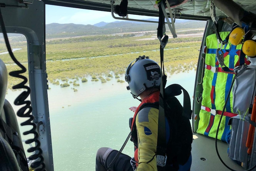 A rescue crew person looks out the open door of a helicopter over marsh land and water.
