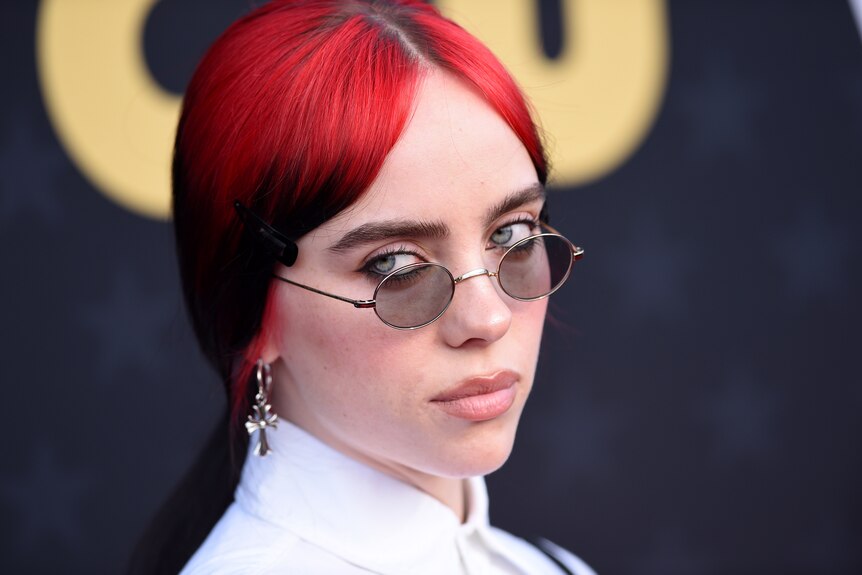 Billie Eilish with bright dyed red hair looks sternly over the tops of little john lennon glasses