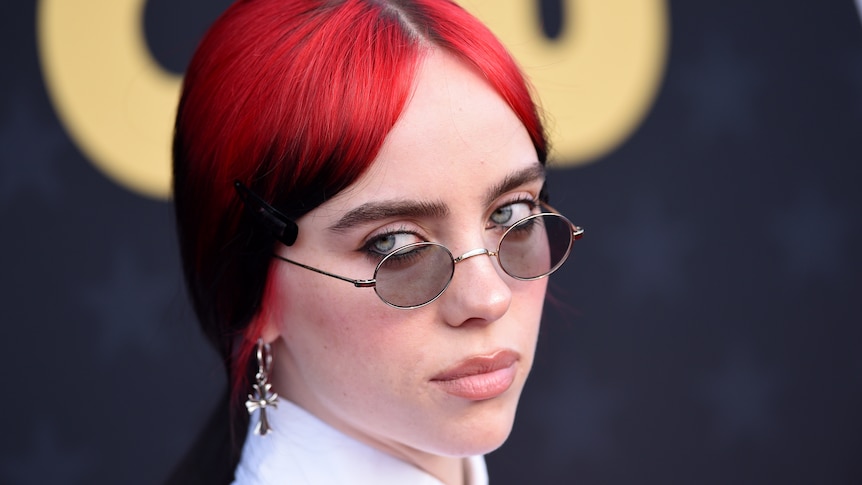 Billie Eilish with bright dyed red hair looks sternly over the tops of little john lennon glasses