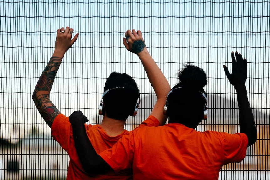 We see the backs of two women in orange t-shirts who look to the outside world through prison bars.