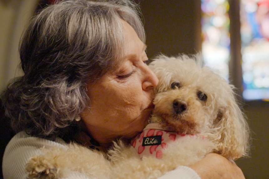 A woman holds a white scruffy dog up to her face, church stained glass windows in background