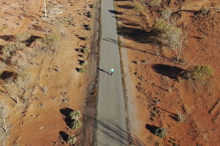 A drone shot of a tiny figure in the middle of a long stretch of outback road.