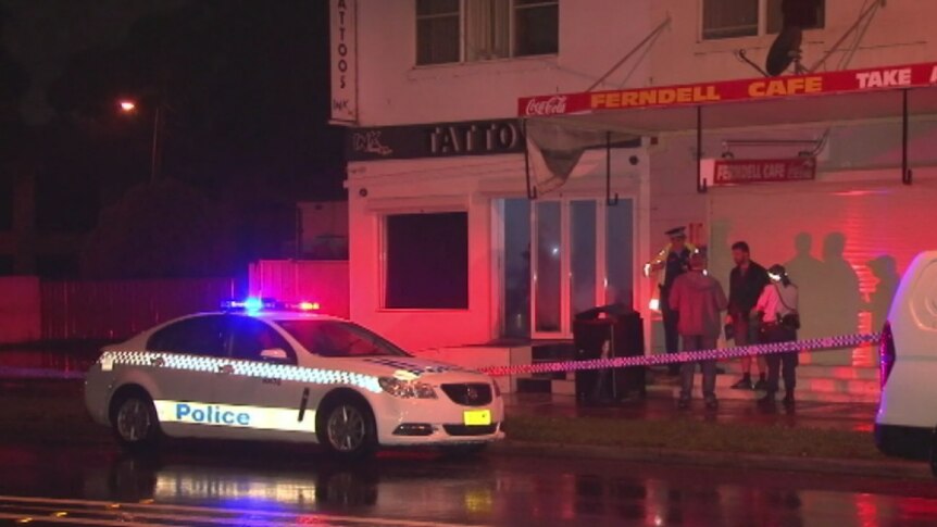Two police officers speak to two men behind police tape outside the tattoo parlour at night
