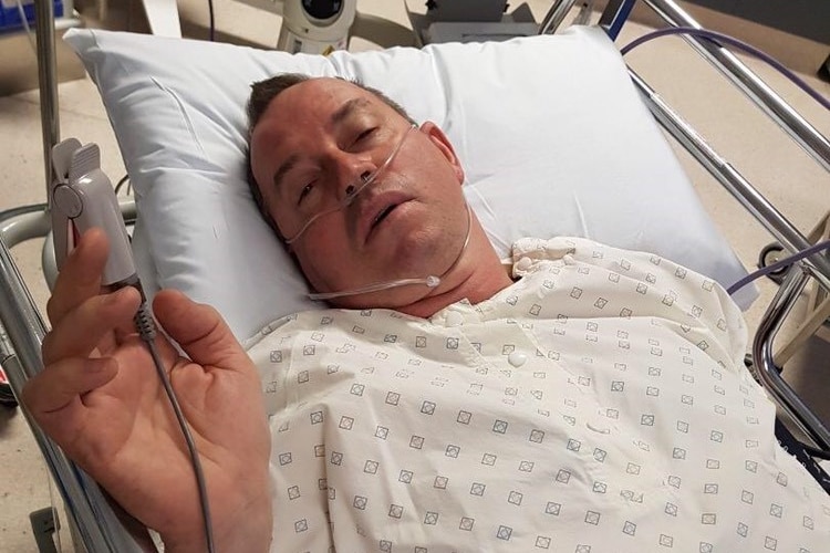 A man lies in a hospital bed with tubes on his face in a hospital gown