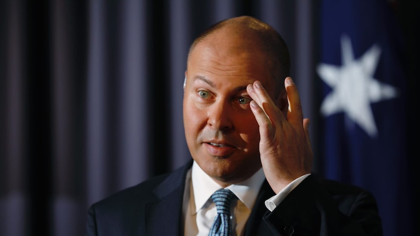 A balding man in a suit stands in front of an Australian flag touching his eyebrow.