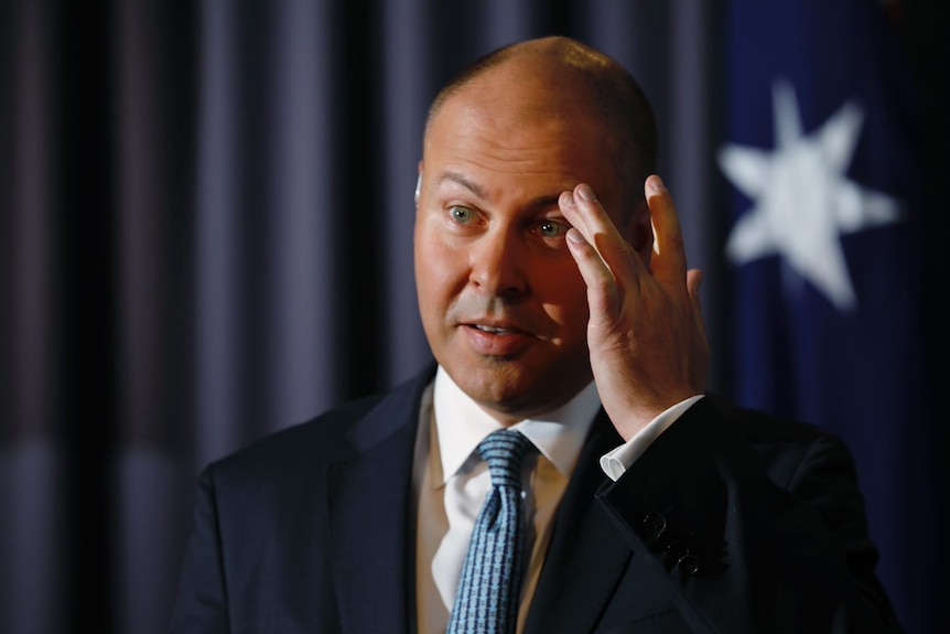 A balding man in a suit stands in front of an Australian flag touching his eyebrow.
