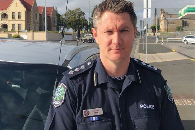 A police officer with a sensible, if slightly rakish haircut, stands in front of a patrol car, squinting in the sun.