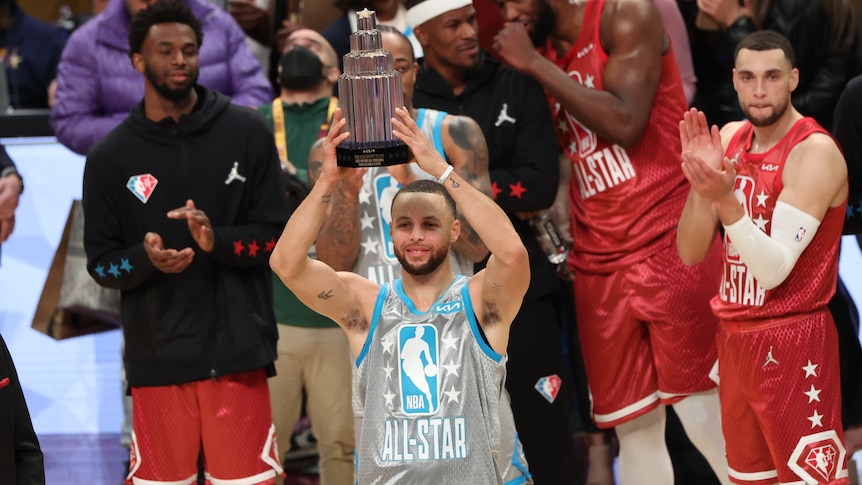 Steph Curry smiles while holding up a large silver trophy. Players applaud him in the background