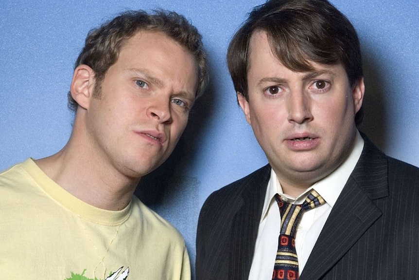 Jeremy and Mark look at the camera in front on a blue wall in this Peep Show promo image.