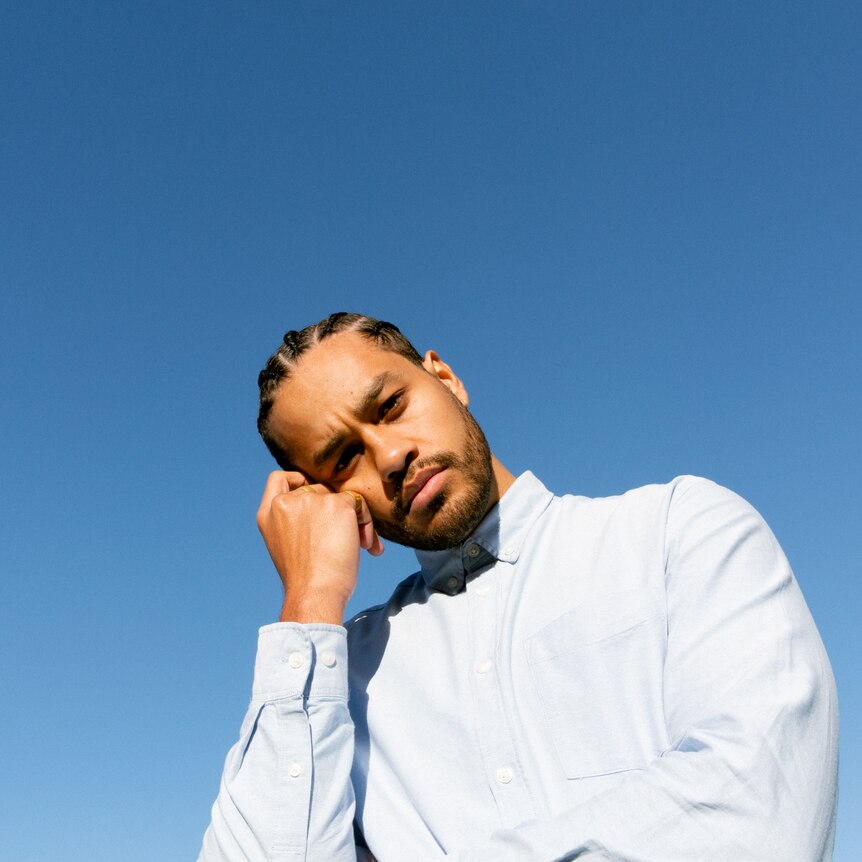 Ziggy Ramo in front of a blue background, wearing a light blue shirt, head resting on hand, cornrows