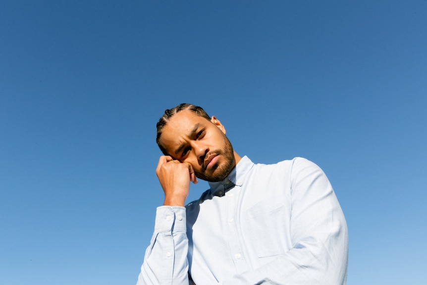 Ziggy Ramo in front of a blue background, wearing a light blue shirt, head resting on hand, cornrows