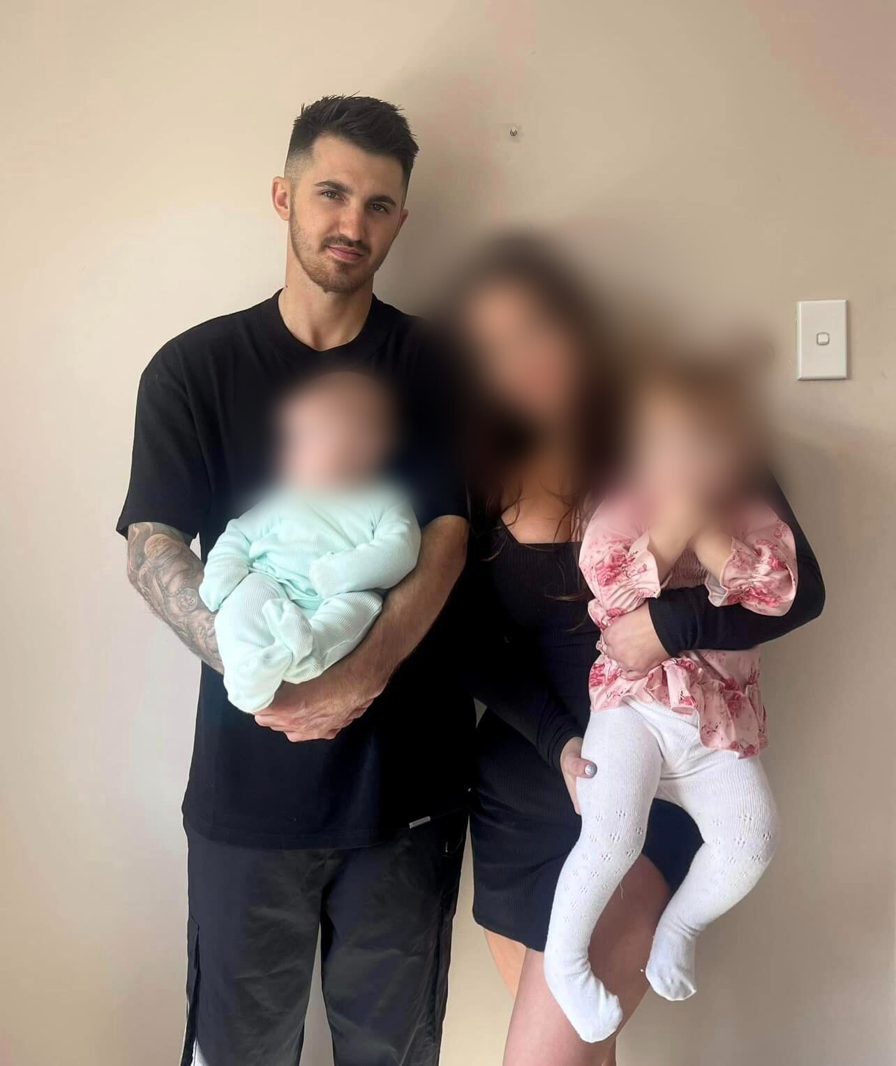 A man in a black shirt surrounded by his family, a woman and two children whose faces have been blurred. 