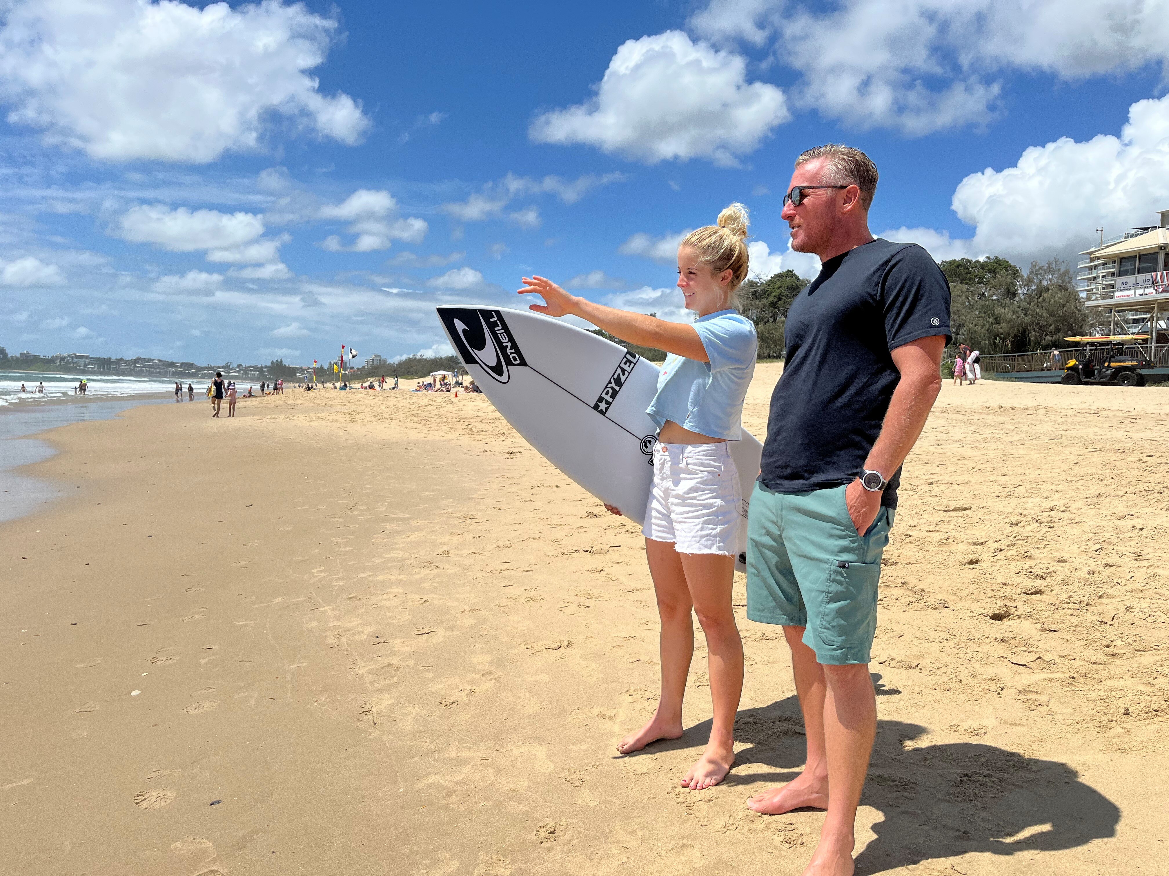Young woman with surfboard pointing at waves with male coach in sunglasses