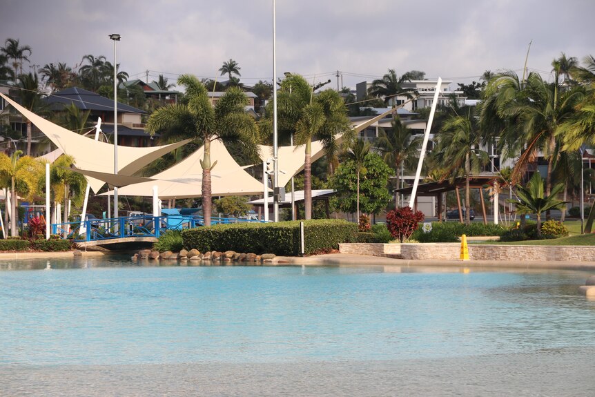 A man-made swimming lagoon with shade sails in the background and aplm trees.