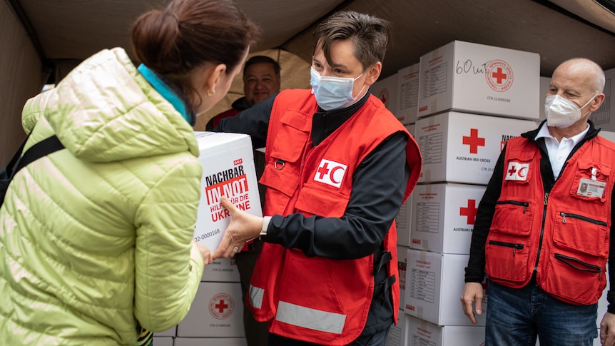 A masked woman in red cross uniform handing over a box