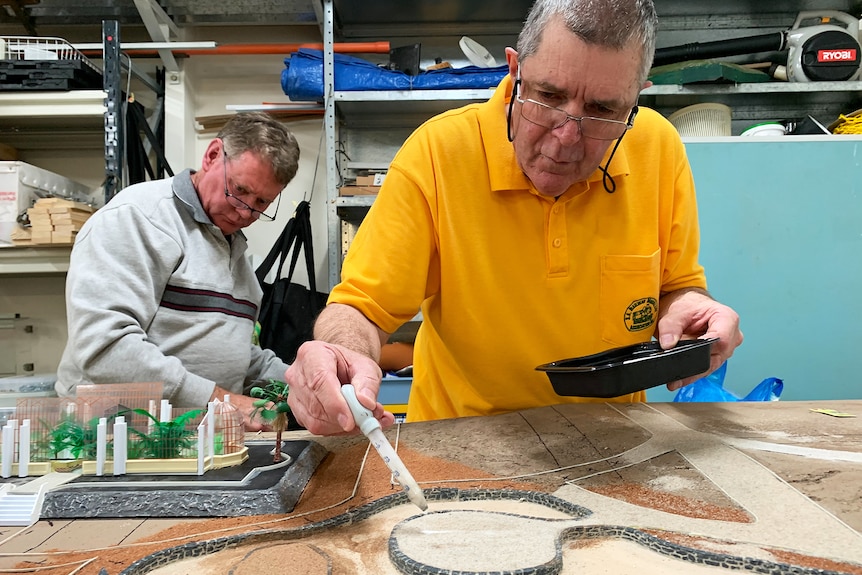 A man in a yellow T-shirt paints a model while another man looks in the background