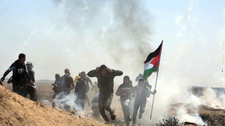 A group of men is seen running for cover, surrounded by tear gas, and one is holding a Palestinian flag.