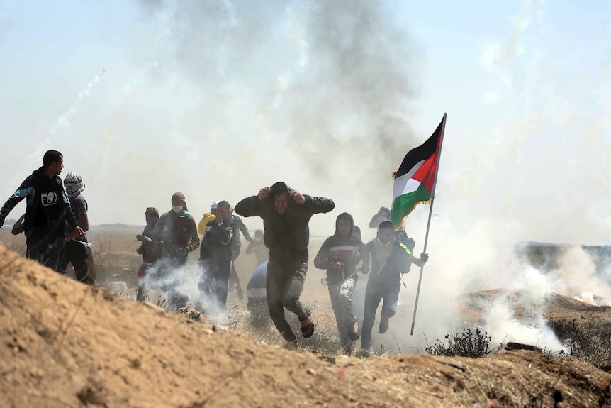 A group of men is seen running for cover, surrounded by tear gas, and one is holding a Palestinian flag.