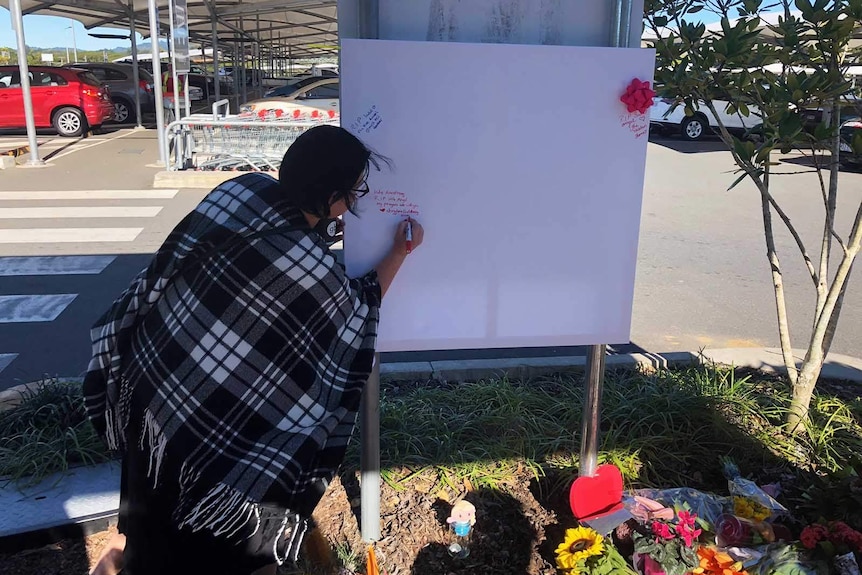 A woman writes on a white card next to flowers at a crossing in a Nambour shopping centre