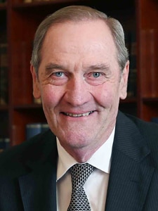 A headshot of an older Caucasian man with green eyes and light silver brown hear smiling at the camera in a suit and tie.