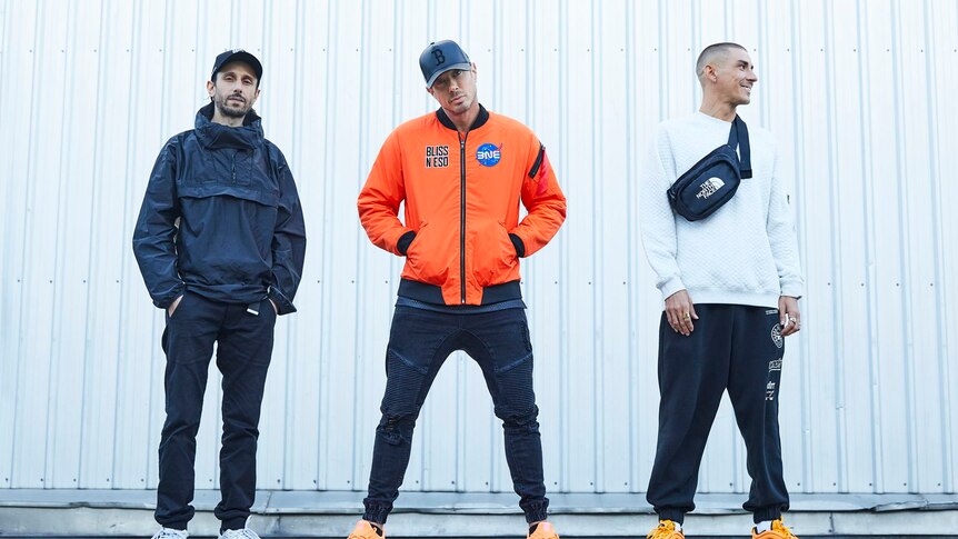 The three members of Bliss N Eso pose in front of a white corrugated fence wearing streetwear and sneakers