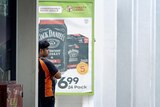 A bottle shop worker leaning against a wall next to a poster promoting Jack Daniels.