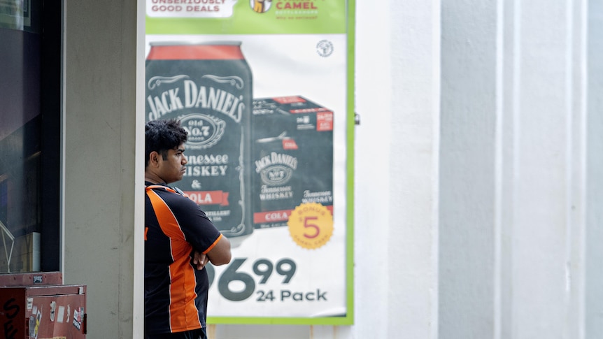 A bottle shop worker leaning against a wall next to a poster promoting Jack Daniels.