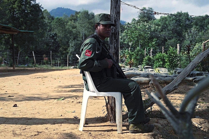 A man in a green uniform with a gun sits on a white plastic chair.