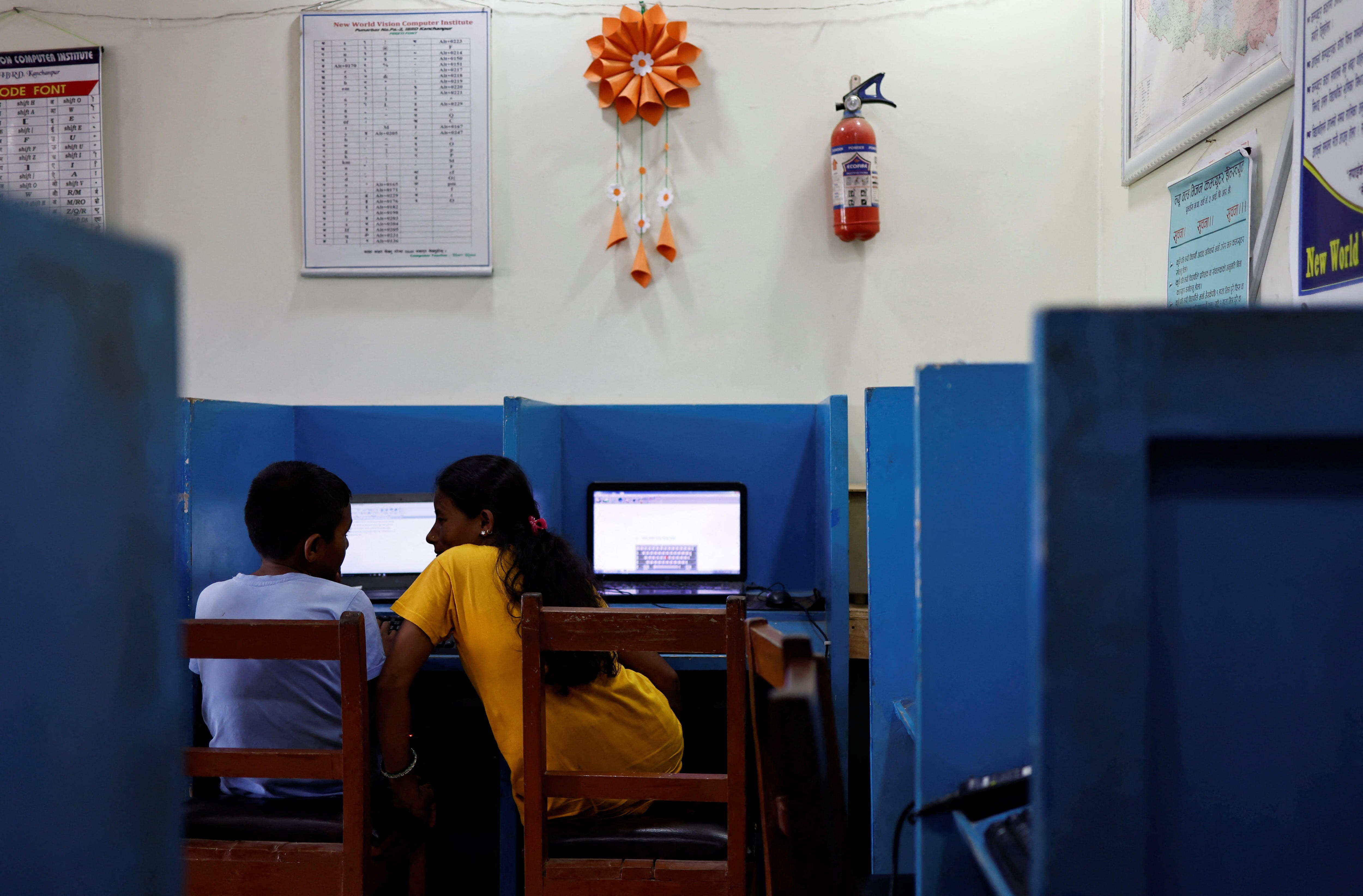 A boy and a woman each sit at laptops in a blue and white classroom. The woman leans over to talk to the boy 