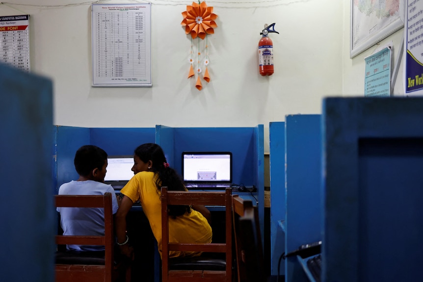 A boy and a woman each sit at laptops in a blue and white classroom. The woman leans over to talk to the boy 