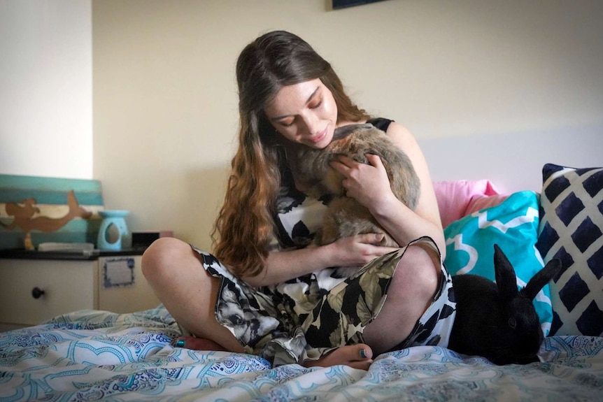A young woman sits on a bed cradling a pet rabbit.