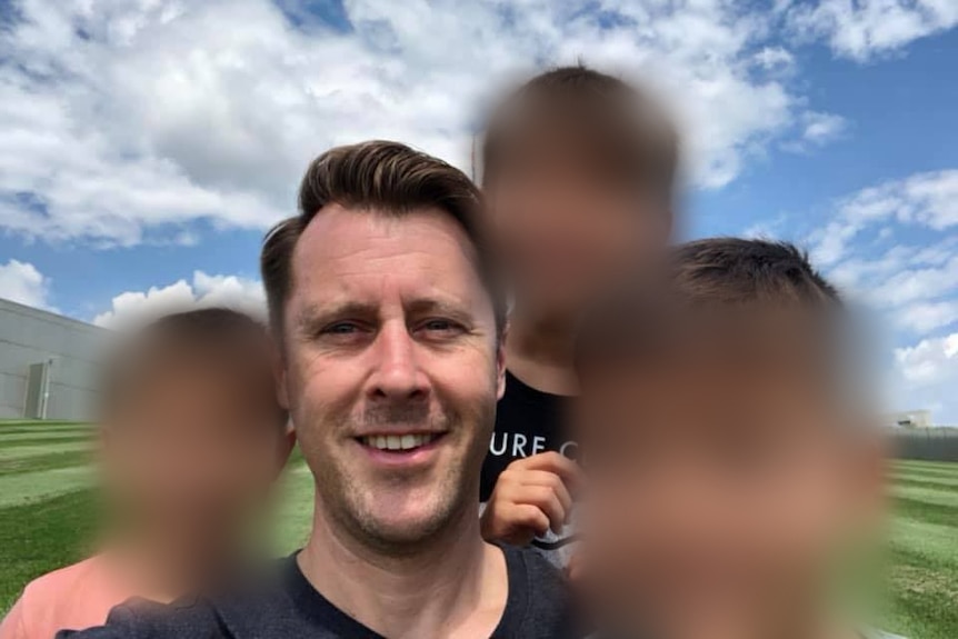 a man smiling in a selfie with three children whose faces are blurred