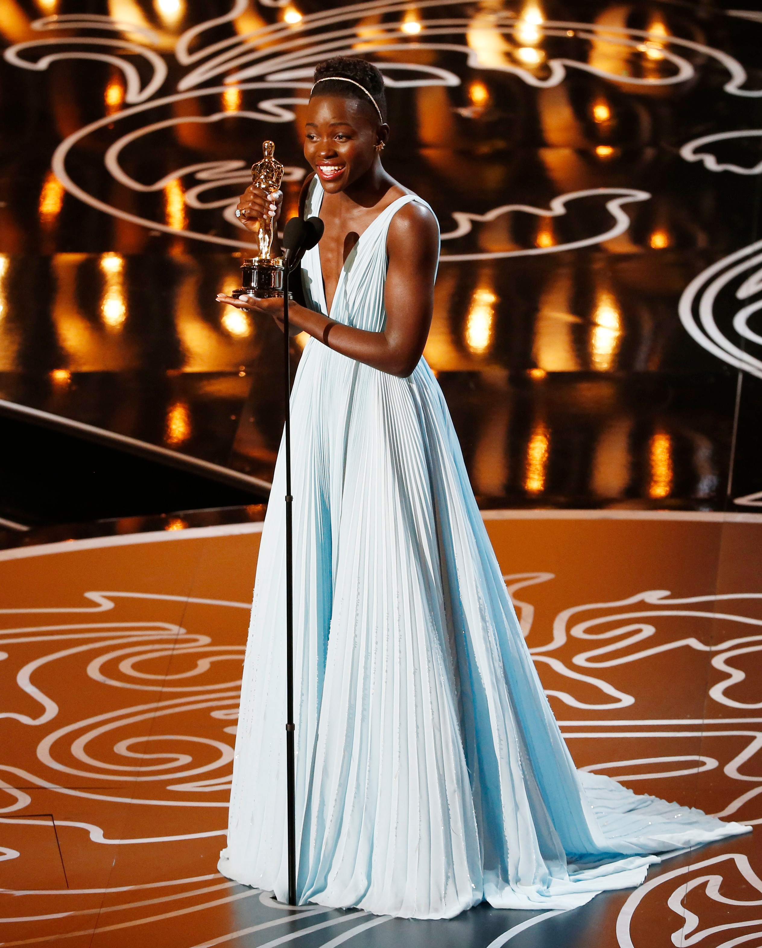 Lupita Nyong'o holding an OScars statue, wearing a powder blue gown with a deep v neck and pleated fabric