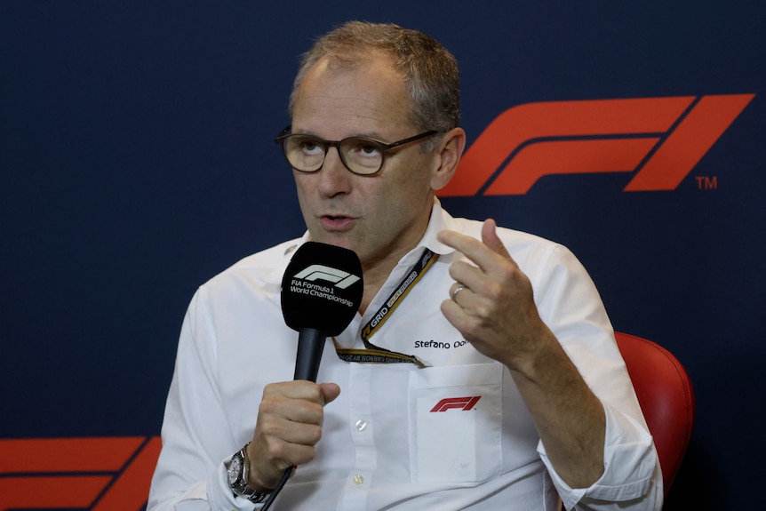 Stefano Domenicali, in a white shirt, holding an F1 branded microphone, speaks at a media conference.